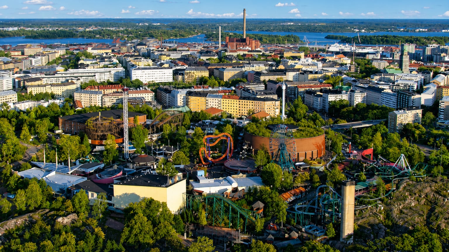 Aerial view of Alppiharju, Helsinki, with Linnanmäki amusement park's roller coasters amid greenery and the city skyline beyond.