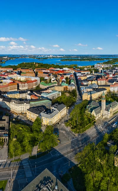 Aerial daytime view over Etu-Töölö, Helsinki, with tree-lined boulevards, dense rooftops and a sea bay beyond.