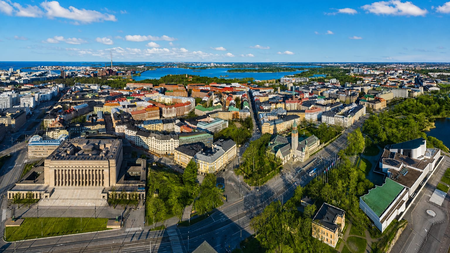 Aerial daytime view over Etu-Töölö, Helsinki, with tree-lined boulevards, dense rooftops and a sea bay beyond.