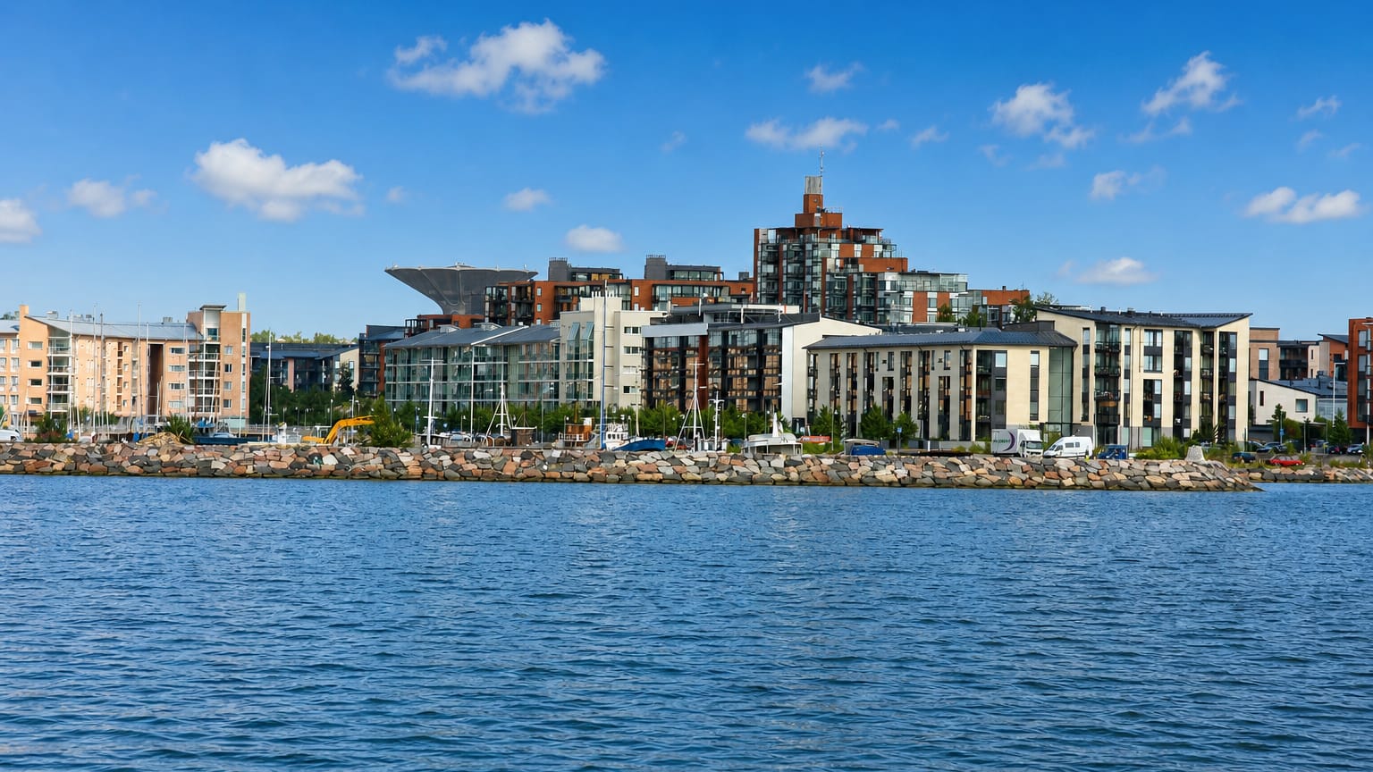 Modern waterfront apartment blocks along the rocky shoreline of Herttoniemenranta, Helsinki, seen across a calm sea bay.