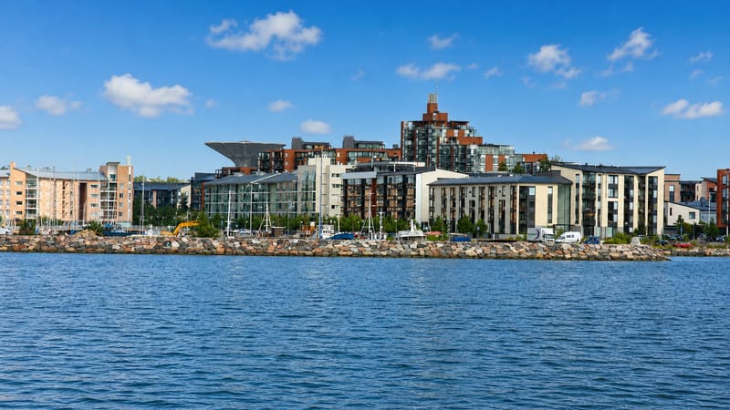 Modern waterfront apartment blocks along the rocky shoreline of Herttoniemenranta, Helsinki, seen across a calm sea bay.