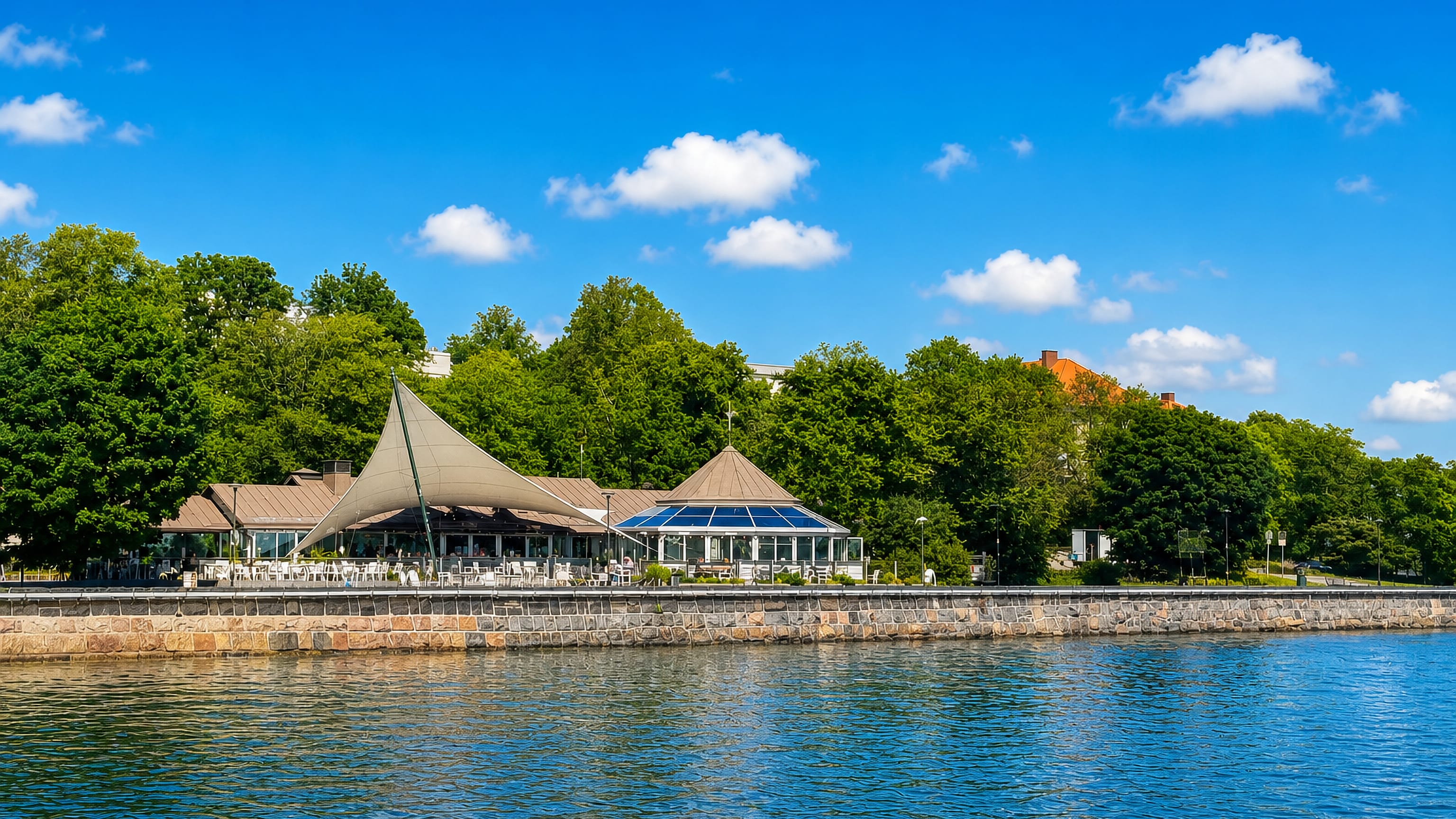 Waterfront cafe pavilion and stone seawall along the shore of Kaivopuisto park in Helsinki on a sunny summer day.