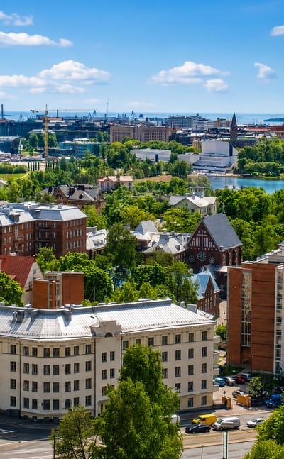 Elevated summer view over Kallio, Helsinki, with old apartment blocks, leafy streets, a bay and the distant city skyline.