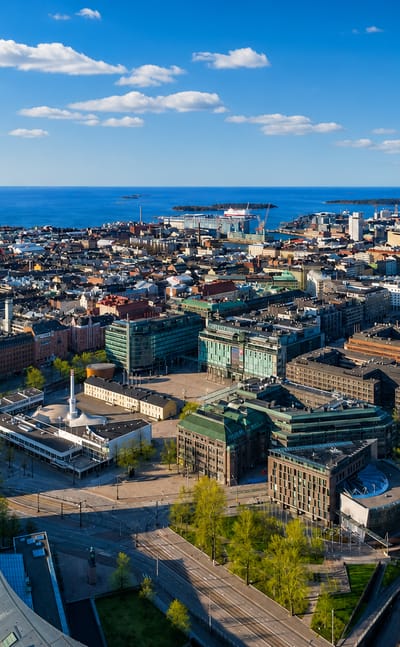 Aerial daytime view over central Helsinki's Kamppi district, dense city blocks reaching the Baltic Sea and offshore islands