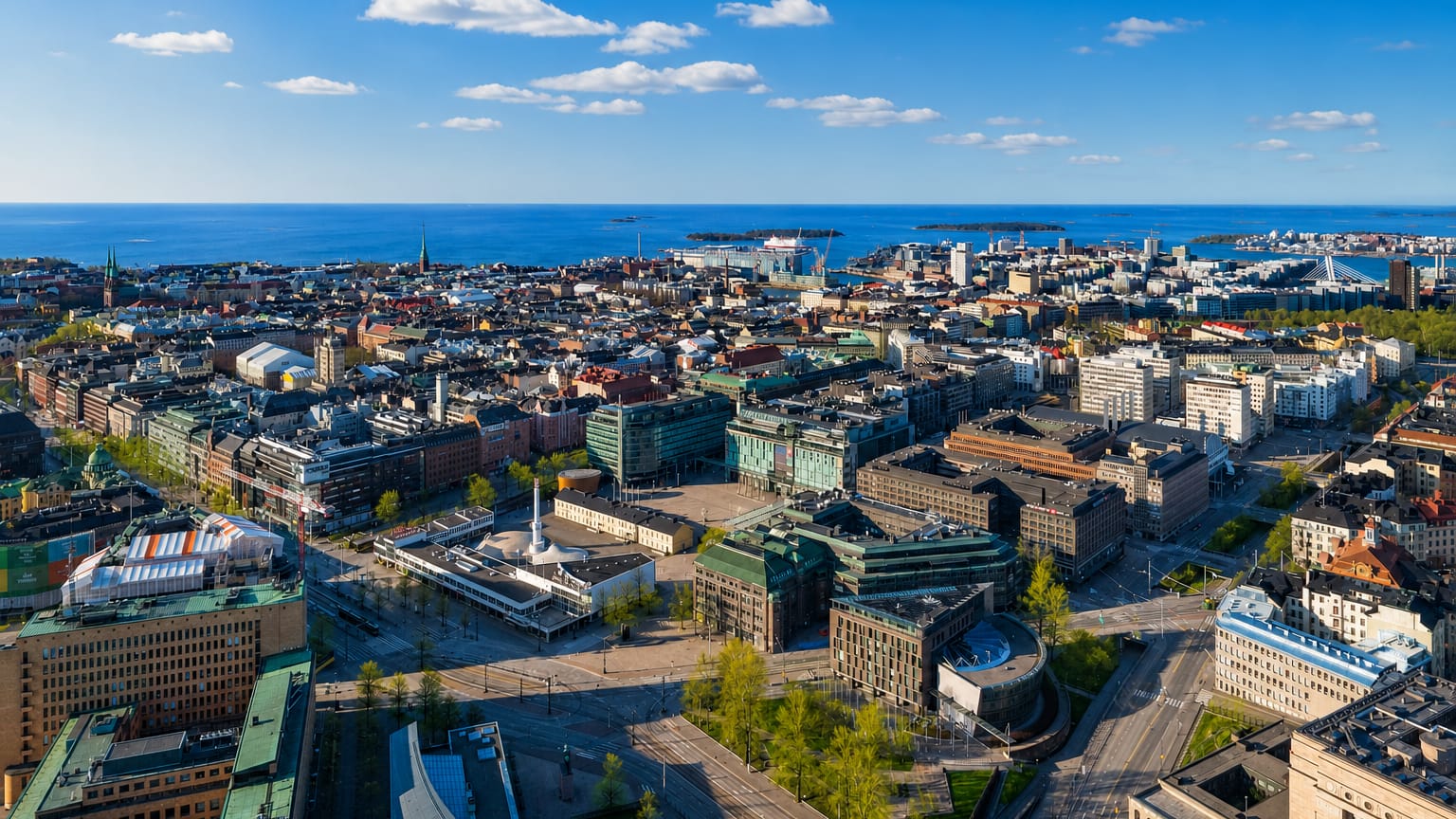 Aerial daytime view over central Helsinki's Kamppi district, dense city blocks reaching the Baltic Sea and offshore islands