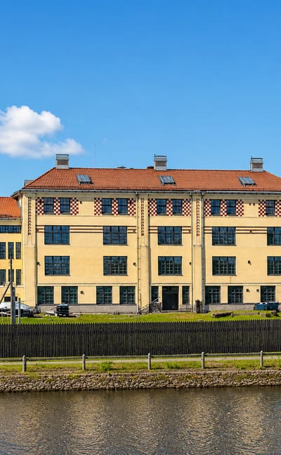 A large yellow three-story building with a red-tiled roof beside a canal under blue sky in Käpylä, Helsinki, Finland.