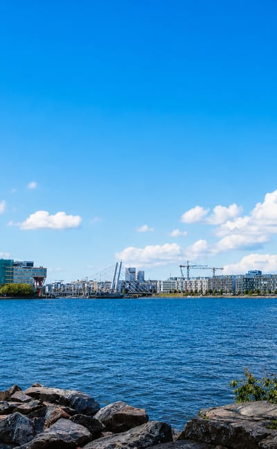 Länsisatama waterfront in Helsinki seen across water, modern mid-rise buildings, construction cranes and a rocky shore.