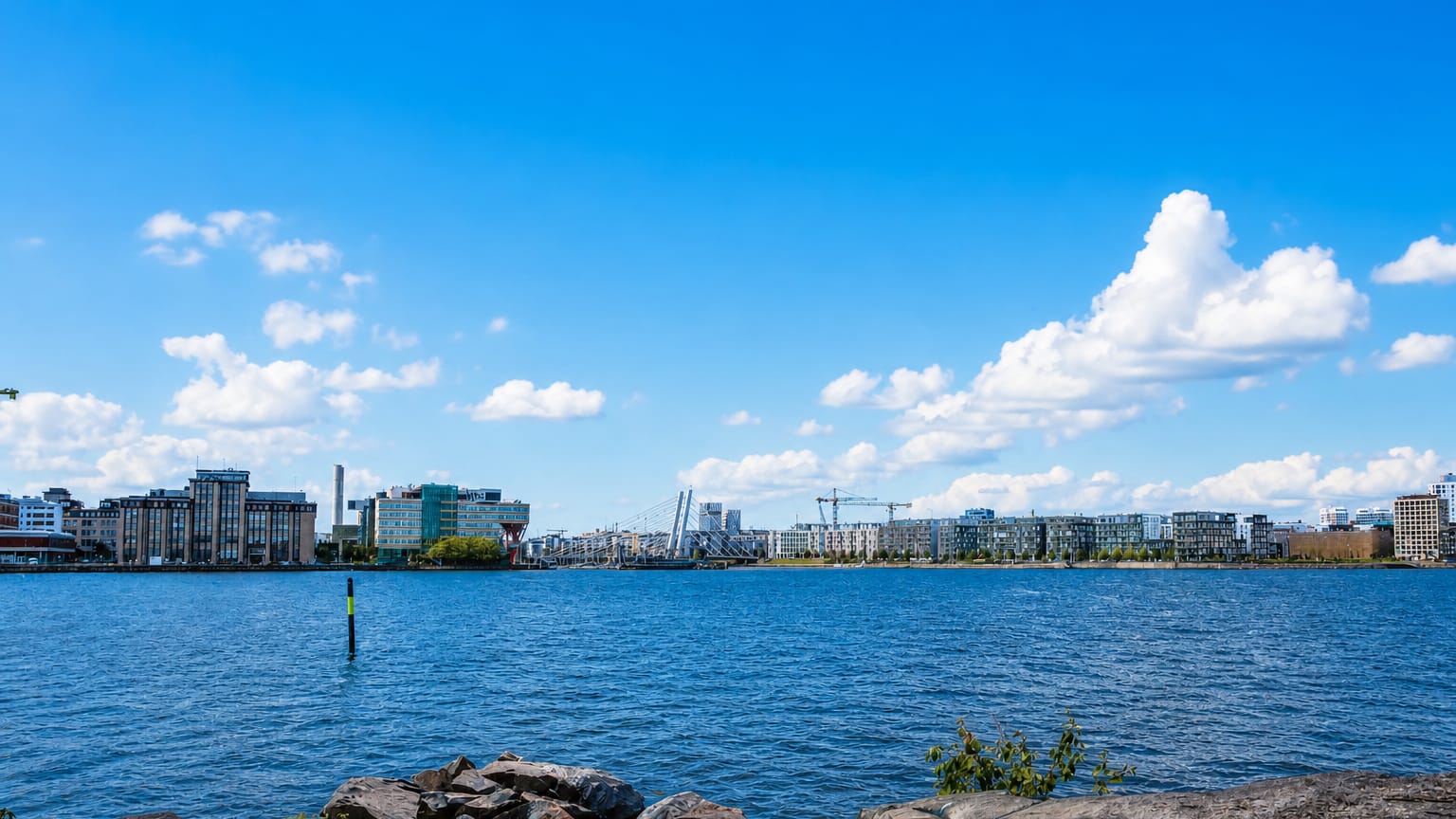 Länsisatama waterfront in Helsinki seen across water, modern mid-rise buildings, construction cranes and a rocky shore.