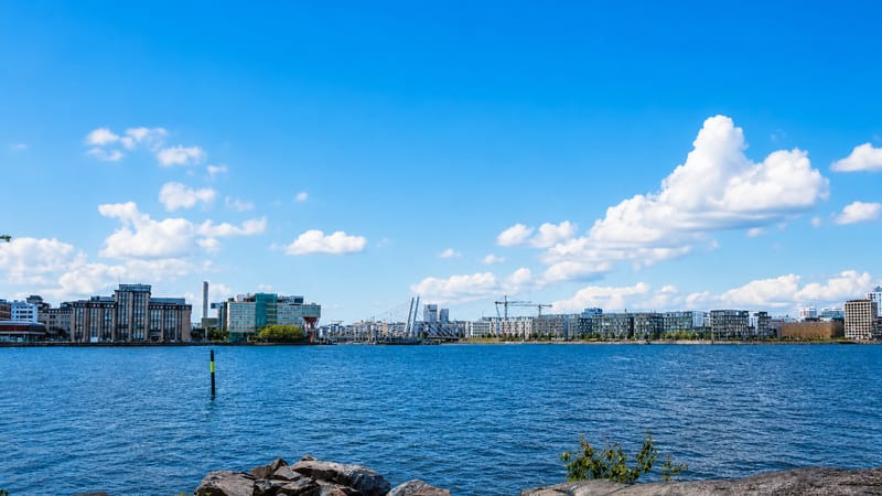 Länsisatama waterfront in Helsinki seen across water, modern mid-rise buildings, construction cranes and a rocky shore.