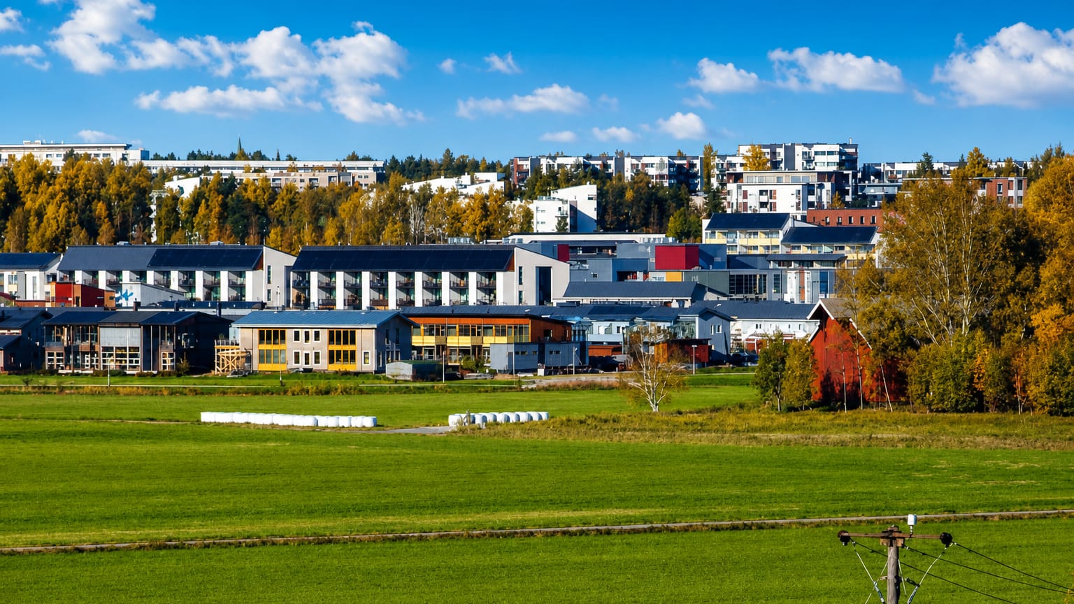 Modern apartment blocks of Latokartano, Helsinki, rising above autumn trees and a green field under a blue sky.
