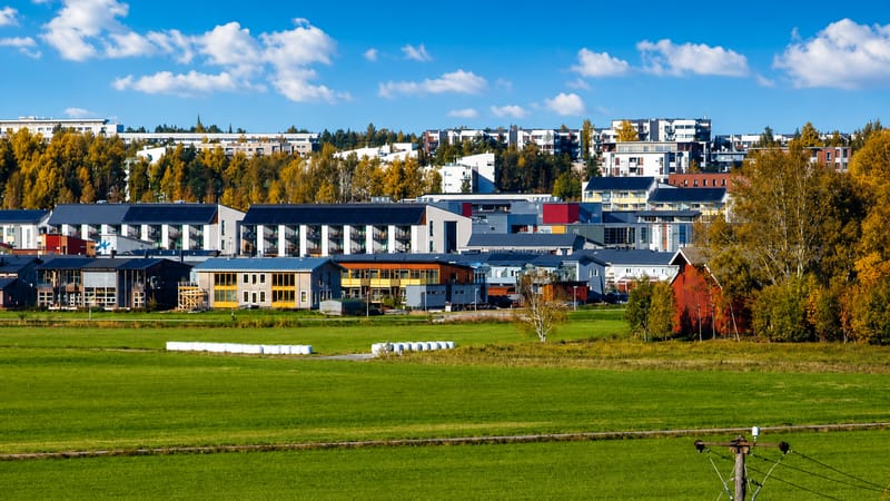 Modern apartment blocks of Latokartano, Helsinki, rising above autumn trees and a green field under a blue sky.