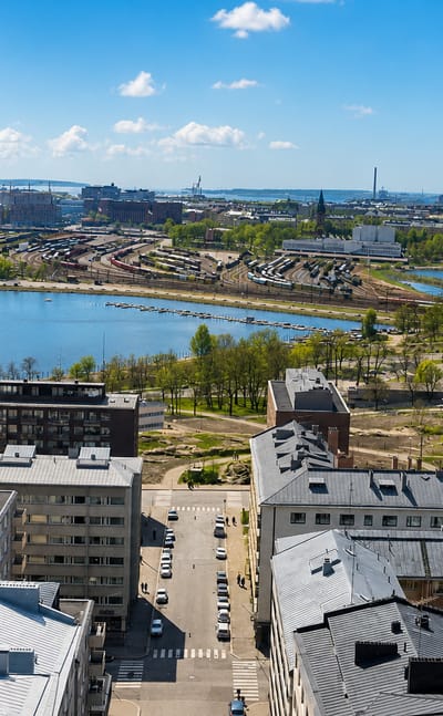 Aerial daytime view over Helsinki's Linjat neighbourhood, dense rooftops overlooking a calm bay and green parkland under a blue sky.