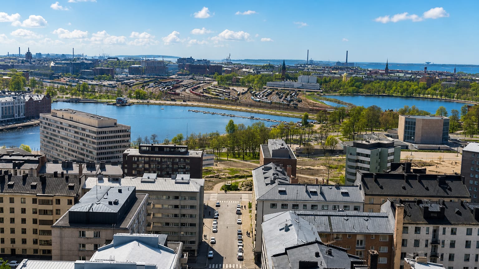 Aerial daytime view over Helsinki's Linjat neighbourhood, dense rooftops overlooking a calm bay and green parkland under a blue sky.