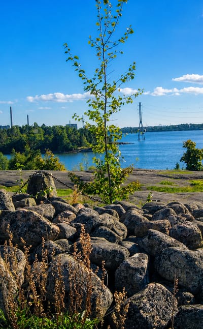Granite boulder shore and calm sea bay near Meilahti, Helsinki, with distant chimneys and a bridge under a sunny summer sky.