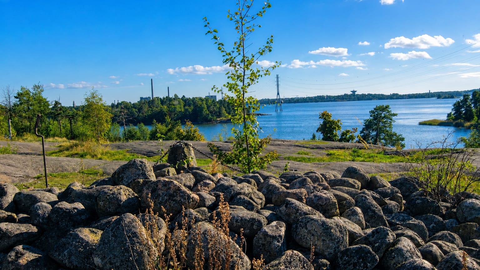 Granite boulder shore and calm sea bay near Meilahti, Helsinki, with distant chimneys and a bridge under a sunny summer sky.