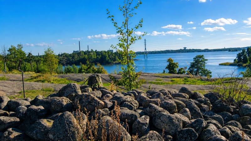 Granite boulder shore and calm sea bay near Meilahti, Helsinki, with distant chimneys and a bridge under a sunny summer sky.