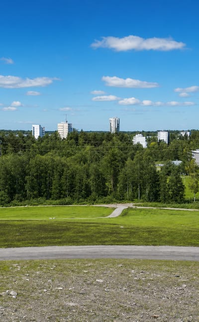 Elevated grassy hill overlooking forest and white residential towers of Myllypuro, Helsinki, under a bright summer sky.
