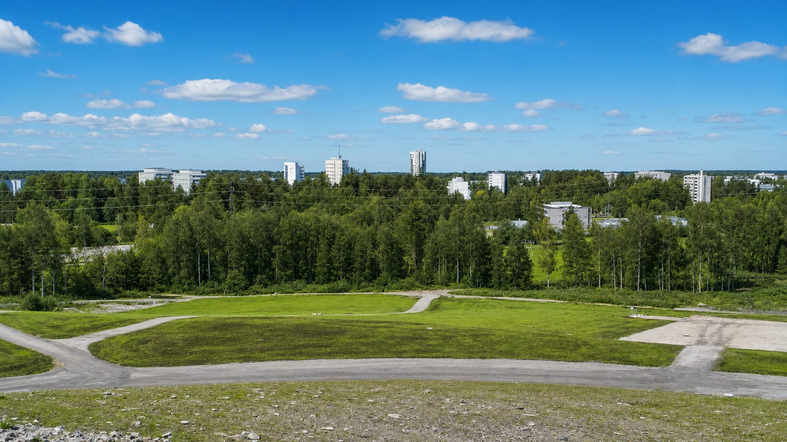 Elevated grassy hill overlooking forest and white residential towers of Myllypuro, Helsinki, under a bright summer sky.