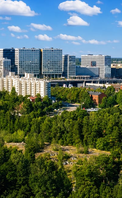Pasila, Helsinki: modern glass office towers and white apartment blocks rising beyond a forested ridge with rail tracks under a blue sky.