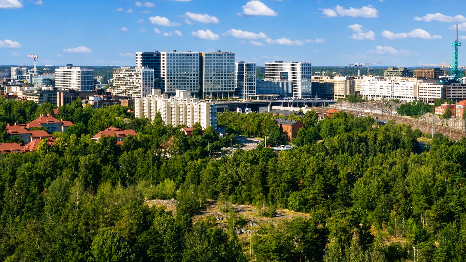 Pasila, Helsinki: modern glass office towers and white apartment blocks rising beyond a forested ridge with rail tracks under a blue sky.