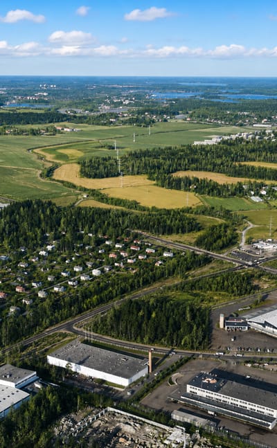 Aerial daytime view over Pitäjänmäki, Helsinki, with industrial buildings, forest, and farmland under a blue cloud-dotted sky.
