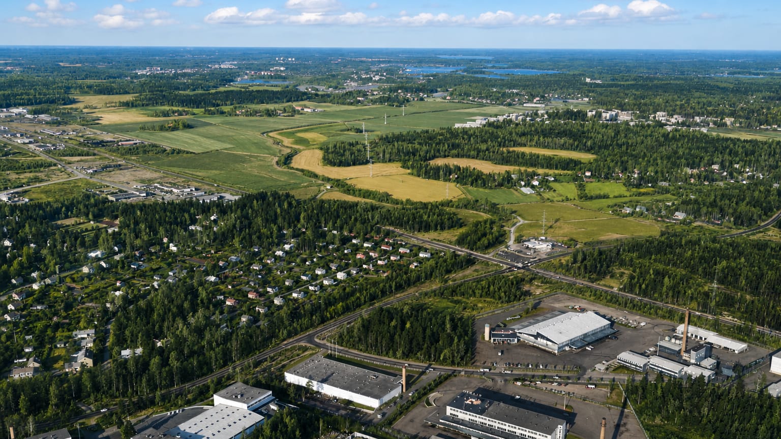Aerial daytime view over Pitäjänmäki, Helsinki, with industrial buildings, forest, and farmland under a blue cloud-dotted sky.