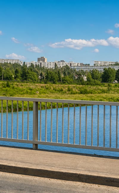 Bridge railing over the Vantaa River in Pukinmäki, Helsinki, with green riverside parkland and hilltop apartment blocks beyond.