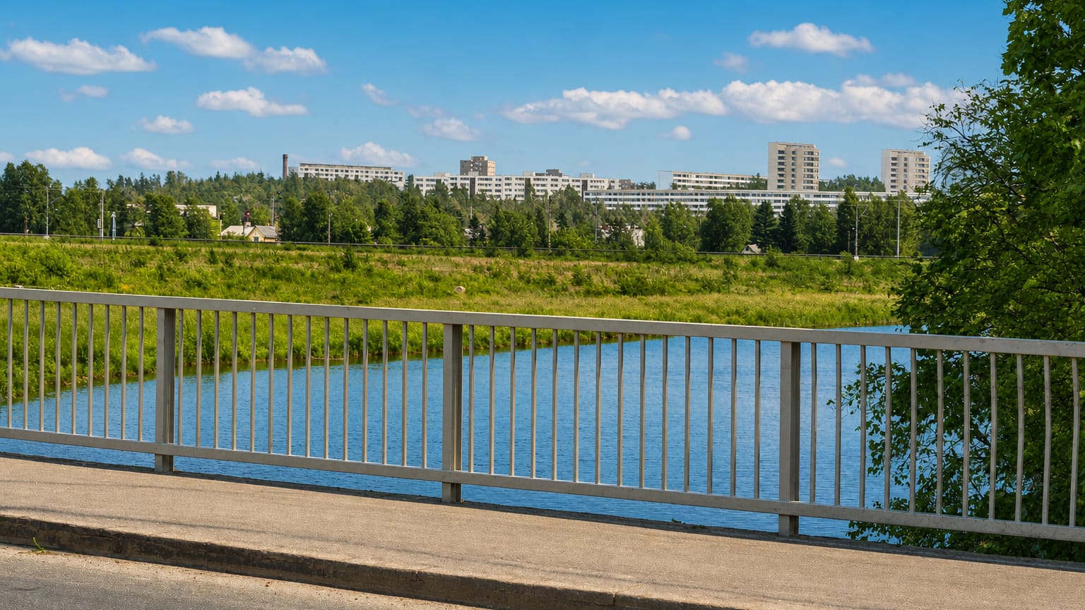 Bridge railing over the Vantaa River in Pukinmäki, Helsinki, with green riverside parkland and hilltop apartment blocks beyond.