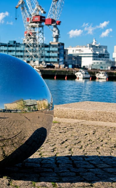 Reflective steel sphere on a cobblestone quay at Hietalahti harbour with red shipyard cranes and moored boats, Punavuori, Helsinki