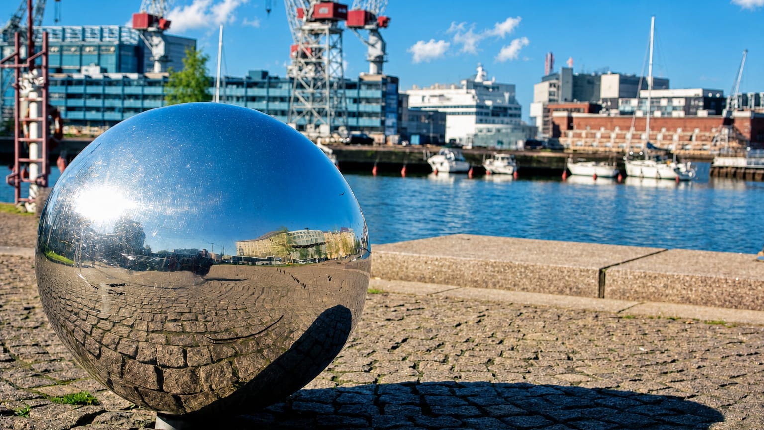 Reflective steel sphere on a cobblestone quay at Hietalahti harbour with red shipyard cranes and moored boats, Punavuori, Helsinki