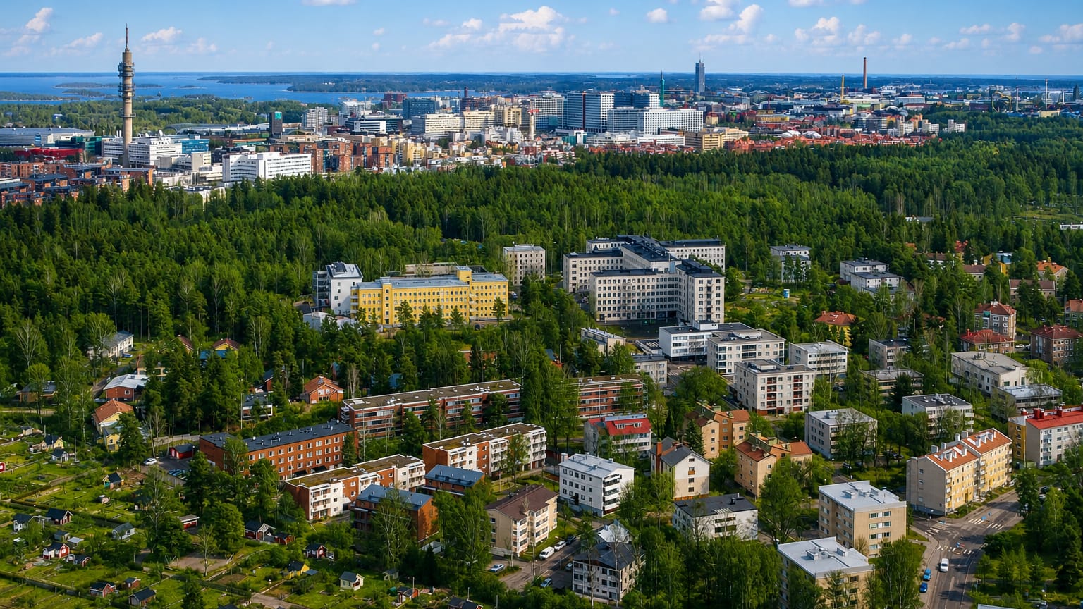 Aerial daytime view of Ruskeasuo's forested apartment blocks and allotment gardens with a lattice tower and Helsinki skyline beyond
