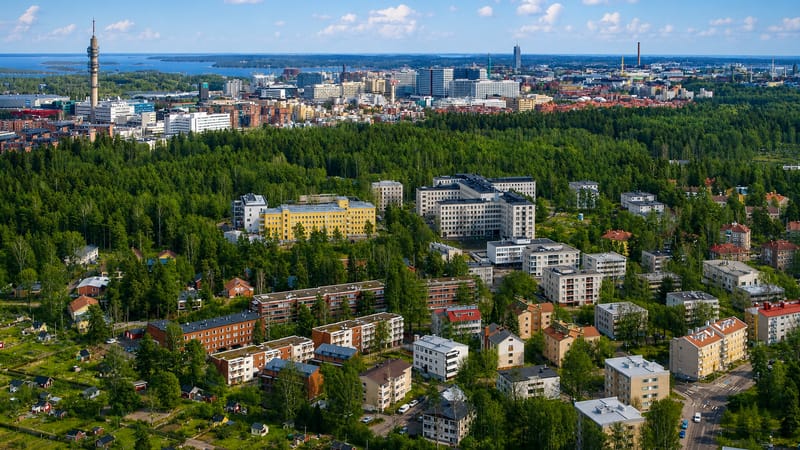 Aerial daytime view of Ruskeasuo's forested apartment blocks and allotment gardens with a lattice tower and Helsinki skyline beyond