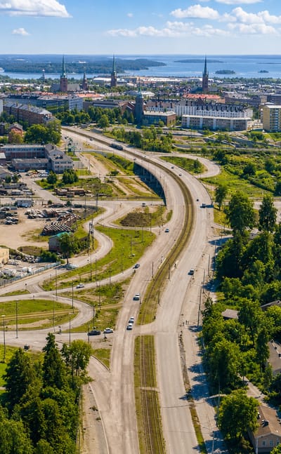 Aerial view of Toukola, Helsinki: a curving road and industrial yards by woodland, with the city skyline and sea beyond.