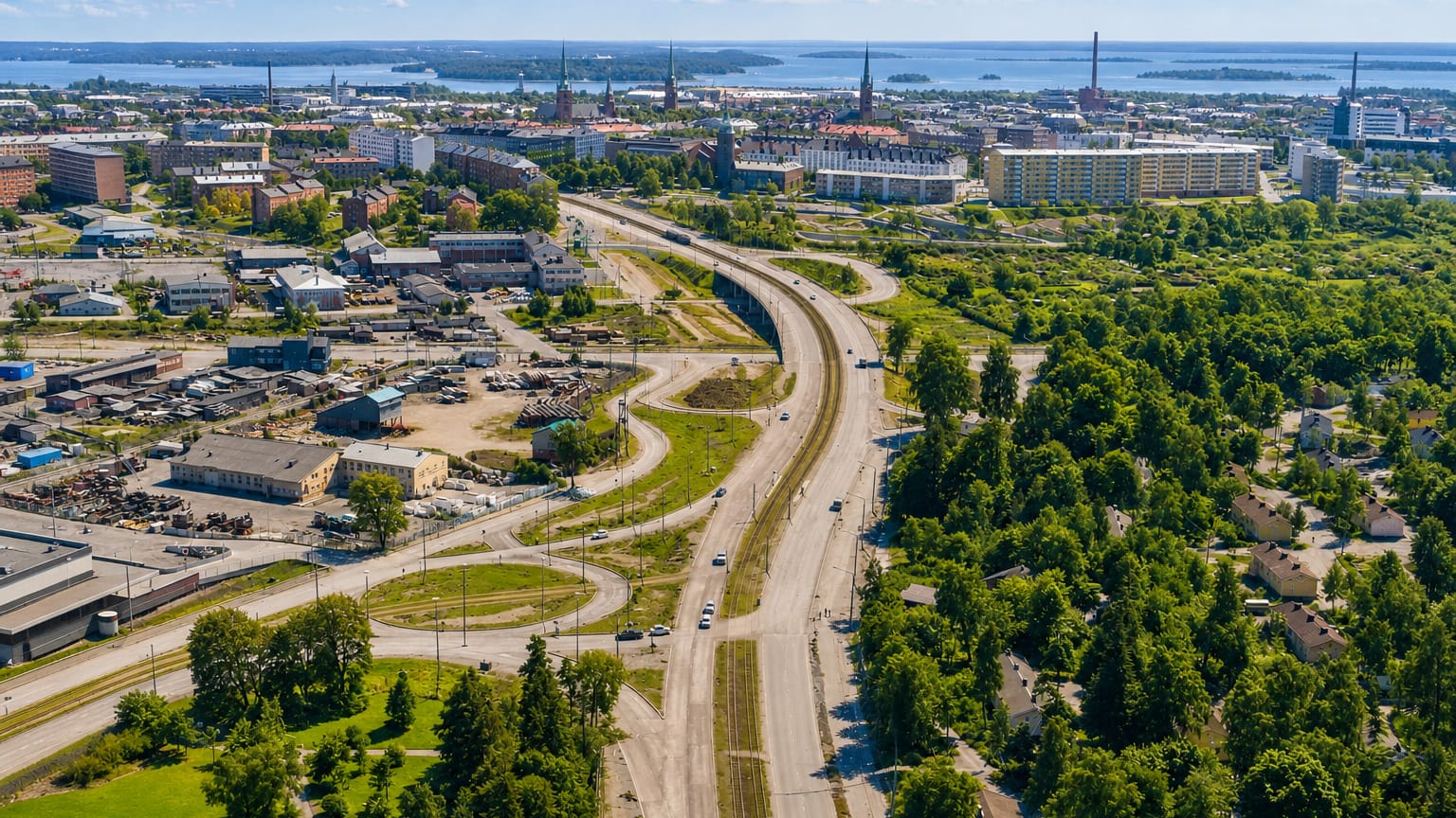 Aerial view of Toukola, Helsinki: a curving road and industrial yards by woodland, with the city skyline and sea beyond.