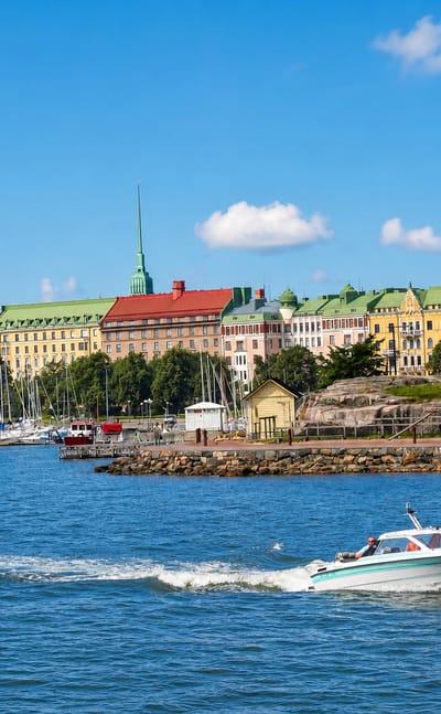 Ullanlinna seafront in Helsinki with pastel 19th-century apartment buildings, a sailboat marina, and a motorboat on the bay.