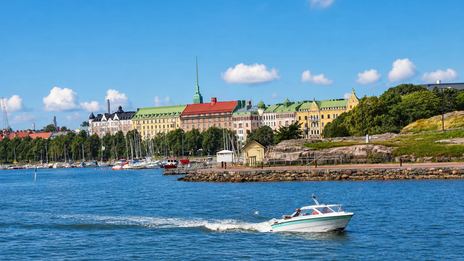 Ullanlinna seafront in Helsinki with pastel 19th-century apartment buildings, a sailboat marina, and a motorboat on the bay.