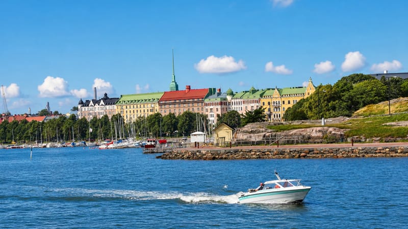 Ullanlinna seafront in Helsinki with pastel 19th-century apartment buildings, a sailboat marina, and a motorboat on the bay.