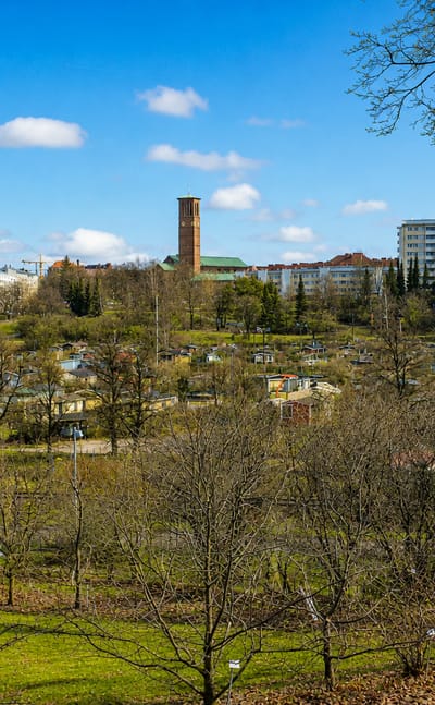 Allotment garden cabins below apartment blocks and a brick tower under a blue spring sky in Vallila, Helsinki, Finland