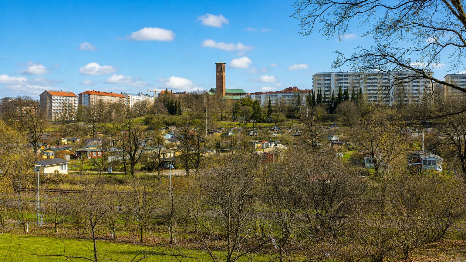 Allotment garden cabins below apartment blocks and a brick tower under a blue spring sky in Vallila, Helsinki, Finland