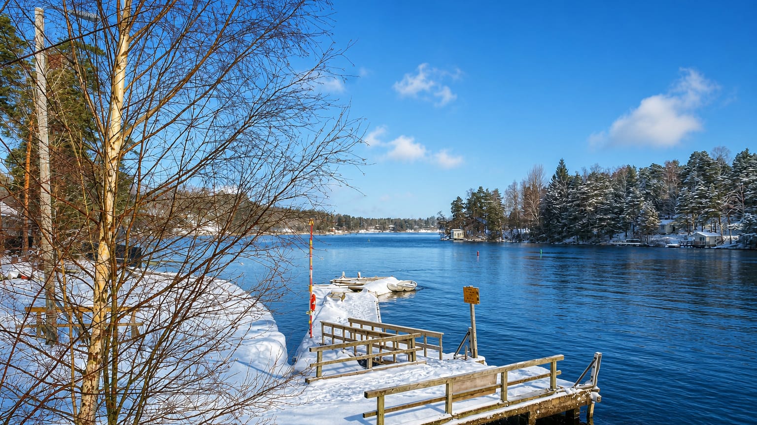 Snow-covered wooden pier reaching into a calm blue sea bay framed by birches and evergreen forest in Yliskylä, Helsinki.