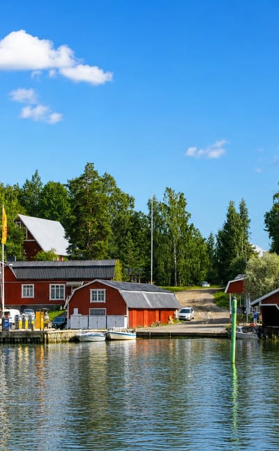 Red wooden boathouses and moored boats along a forested waterfront in Ingå, Finland, under a bright summer sky.