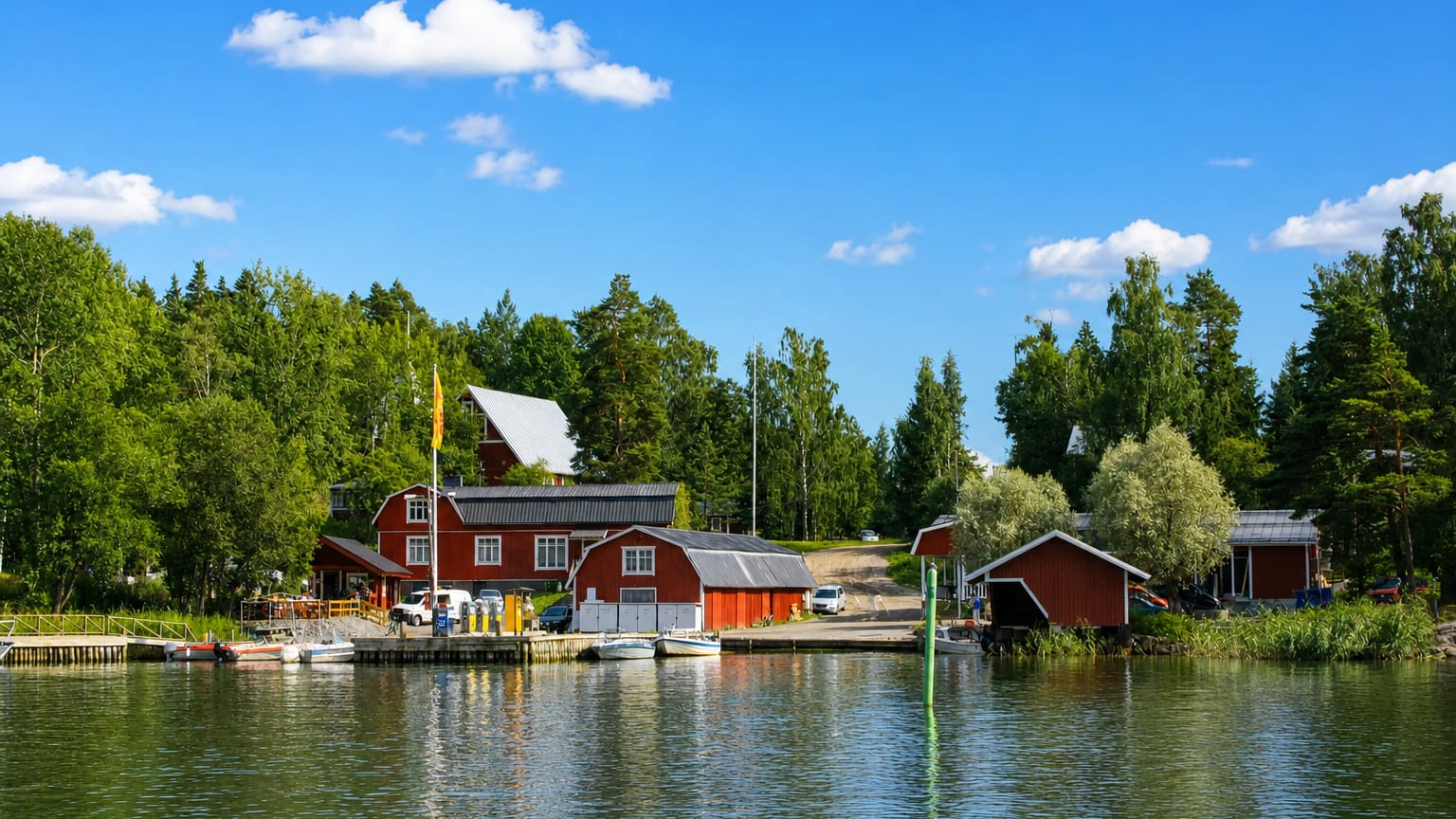 Red wooden boathouses and moored boats along a forested waterfront in Ingå, Finland, under a bright summer sky.