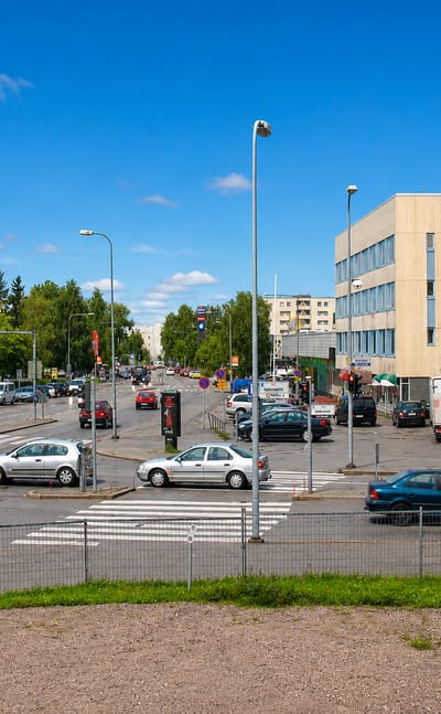 Cars parked around a crosswalk in Jaervenpaeae town centre, lined with low shops and offices under a blue sky.