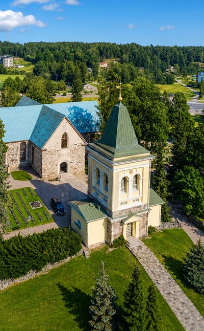 Aerial view of Kirkkonummi's medieval grey stone church and yellow bell tower among trees, with town apartment blocks beyond
