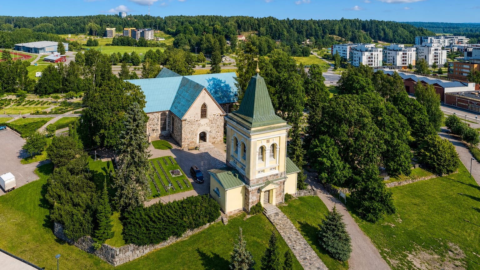 Aerial view of Kirkkonummi's medieval grey stone church and yellow bell tower among trees, with town apartment blocks beyond