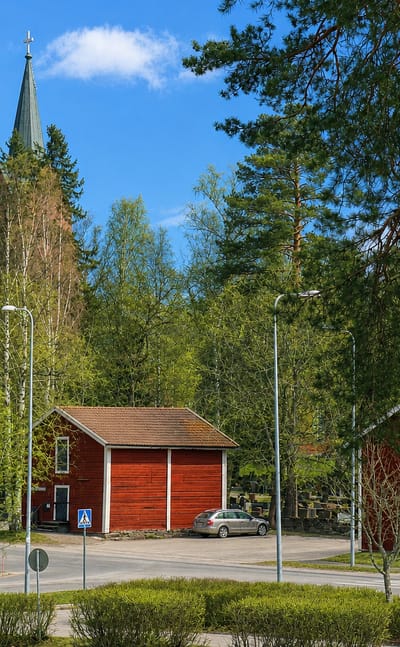 Mäntsälä town centre with a tall wooden church spire above green trees, traditional red storehouses and a quiet road junction