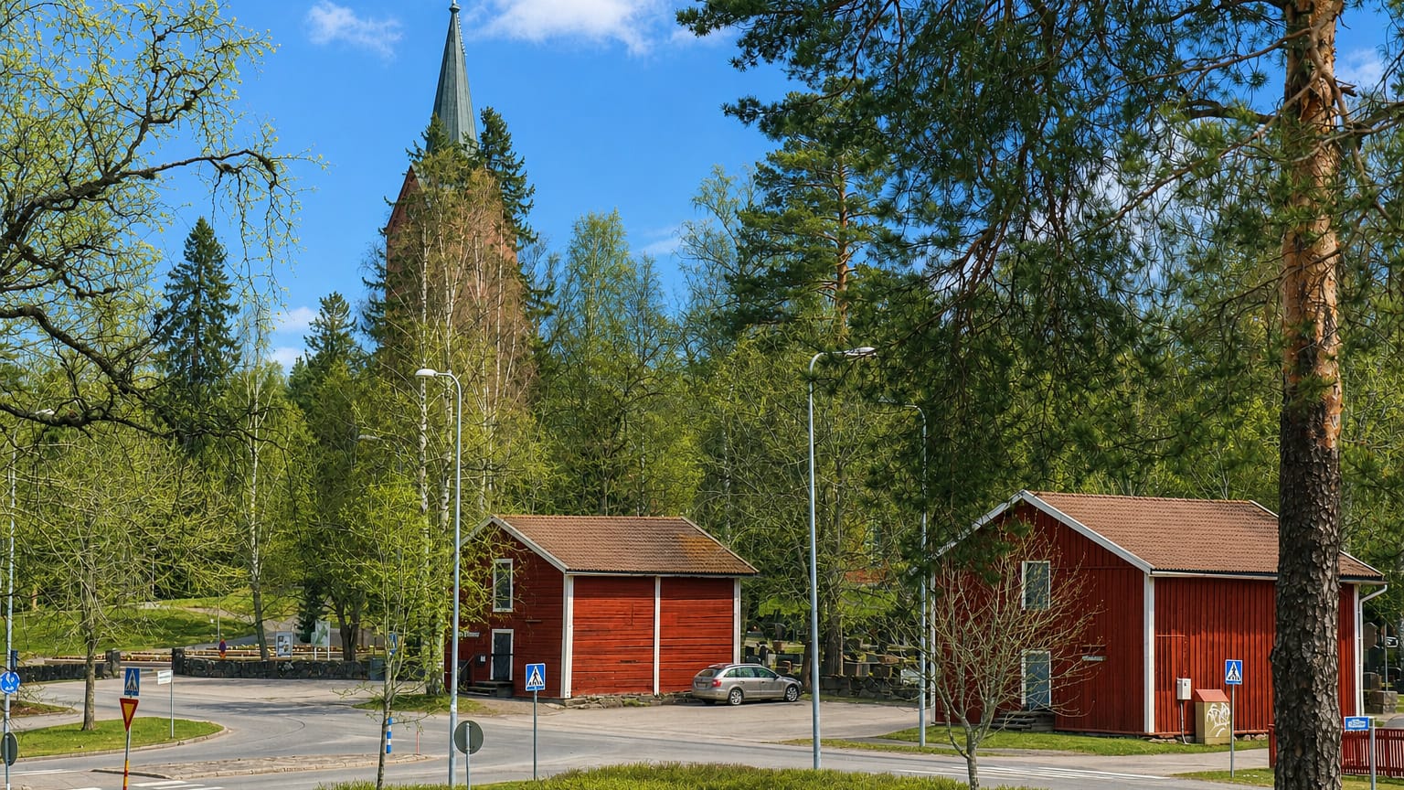 Mäntsälä town centre with a tall wooden church spire above green trees, traditional red storehouses and a quiet road junction