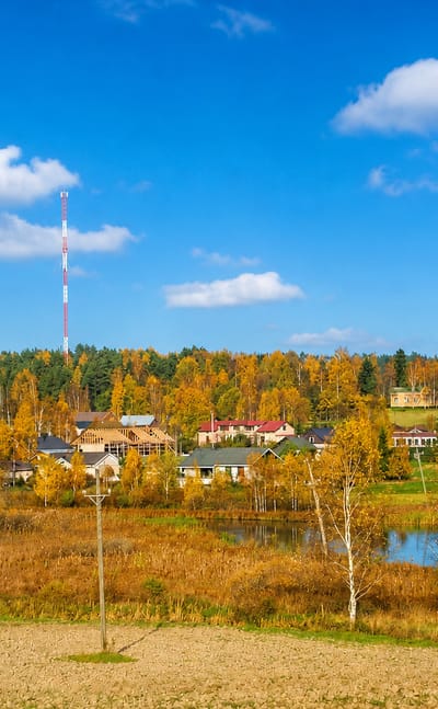 Myrskylä village in autumn, Finland — golden birches, a small lake, low houses and a striped radio tower under blue sky.