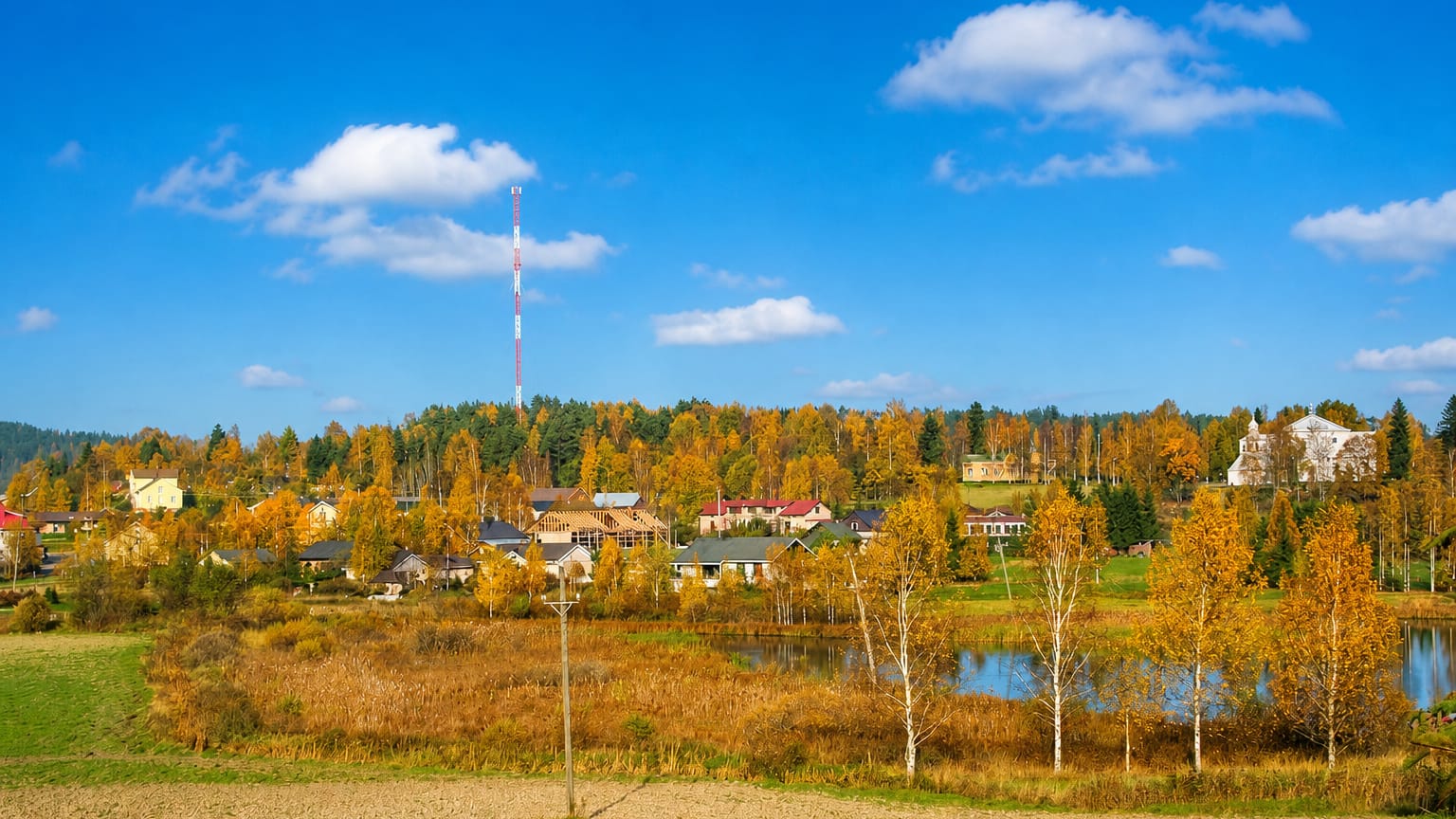 Myrskylä village in autumn, Finland — golden birches, a small lake, low houses and a striped radio tower under blue sky.
