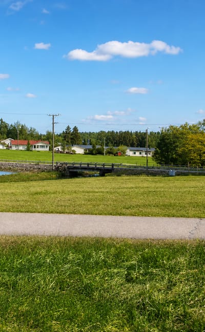 Grassy meadow and footpath by a small pond with a footbridge and red-roofed wooden houses in rural Nurmijärvi, Finland